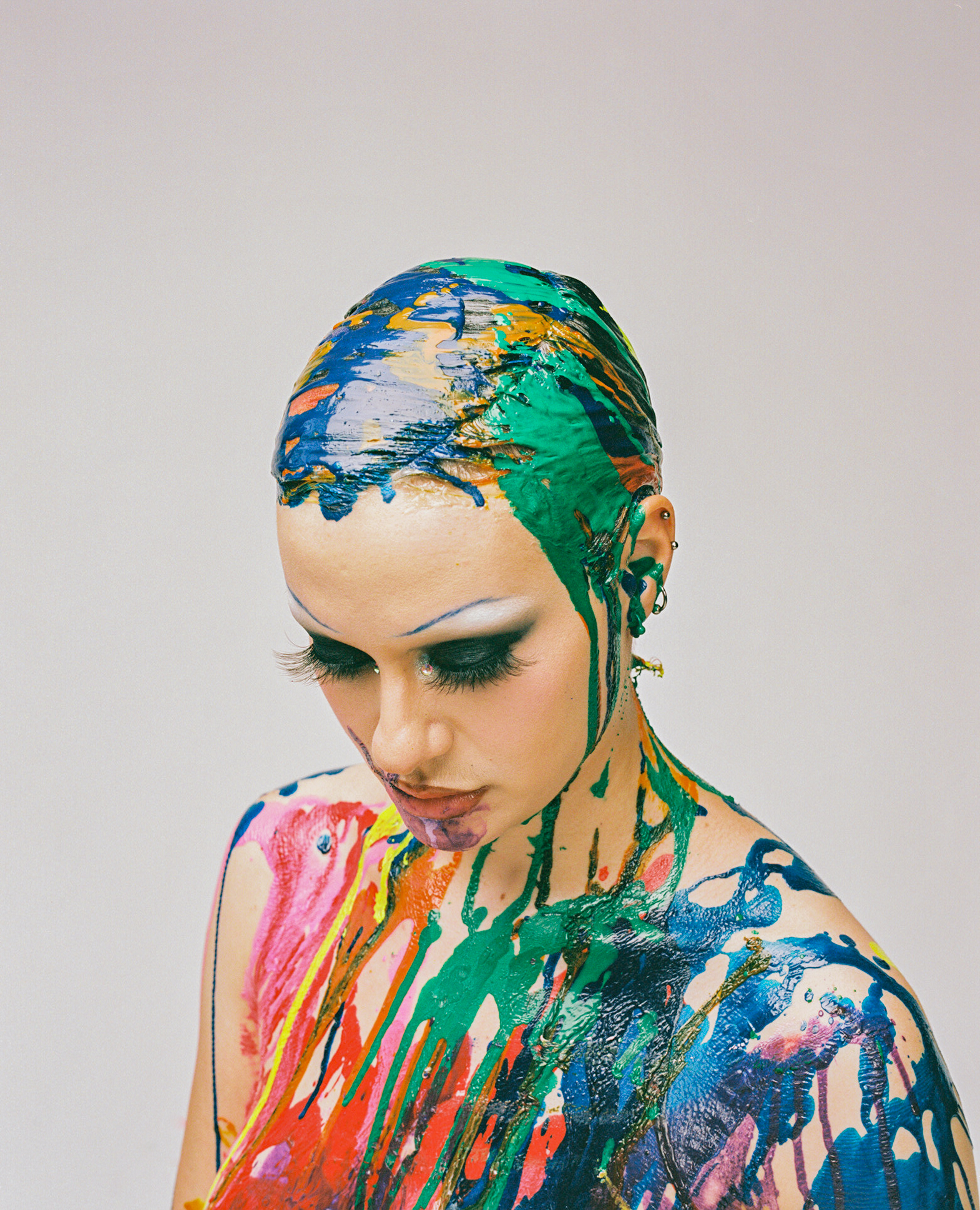 A person with strikingly thin blue eyebrows, deep grey eyeshadow, and extra long eyelashes looks down, their head and upper body covered in colorful, dripping paint against a plain background.